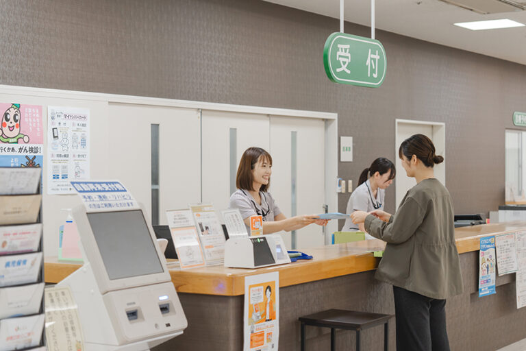 受付カウンターでスタッフが来訪者対応している様子。案内表示と受付システムが見える。